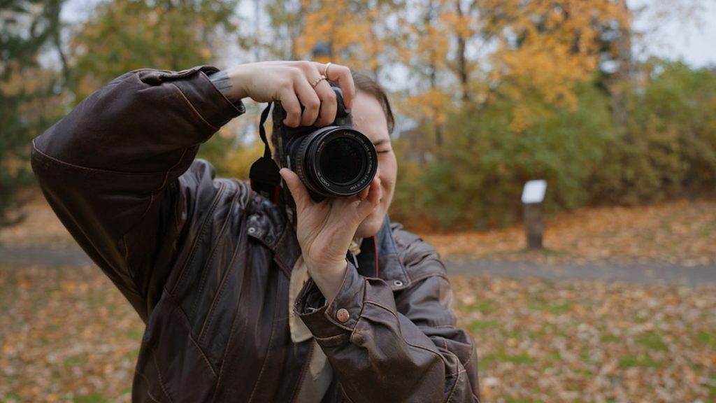 Nuori henkilö ottaa kuvaa järjes­tel­mä­ka­me­ralla, objek­tiivi osoittaa suoraan eteenpäin. Taustalla syksyinen puisto.