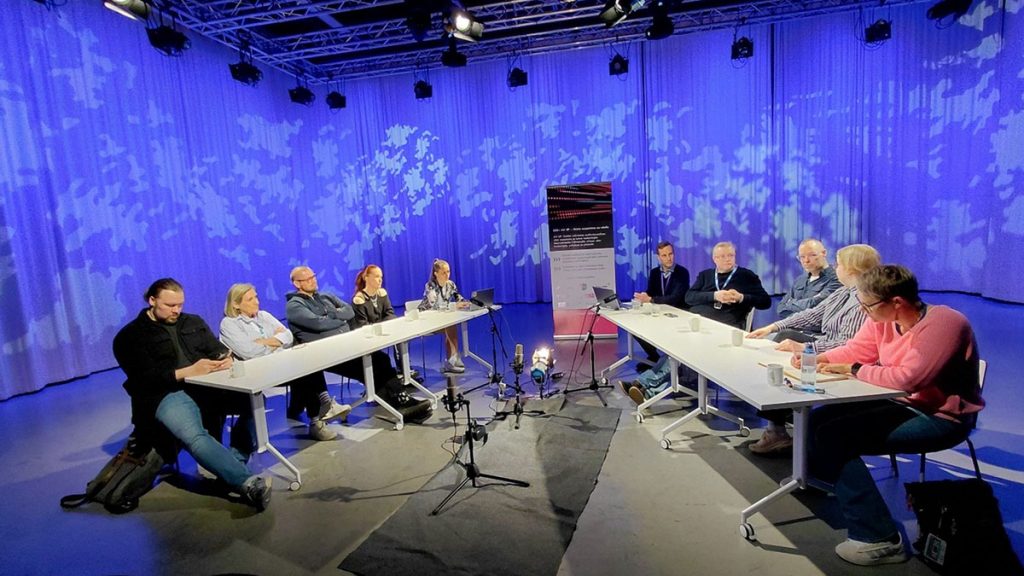 Tio paneldeltagare sitter vid bord i en studio med blå bakgrund.
