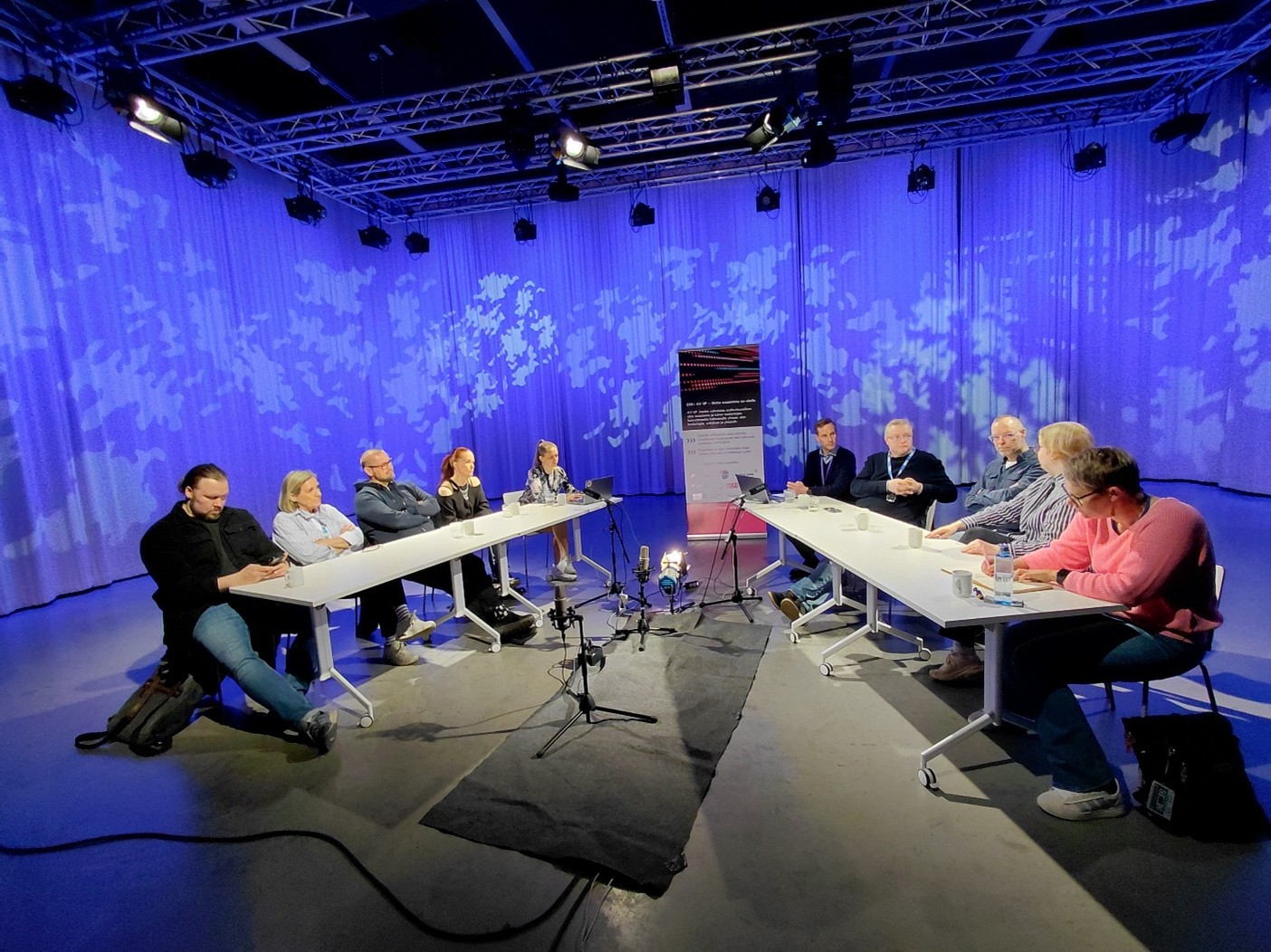 Kymmenen panee­li­kes­kus­te­lijaa istumassa pöytien ääressä sini­taus­tai­sessa studio­ti­lassa.