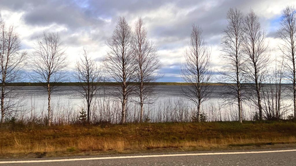 Järven rannalla kasvaa rivi koivuja, vastarannalla näkyy tuntureita. Etualalla on kaistale maantietä, joka kulkee järven vieressä.